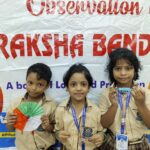 Three schoolgirls in beige uniforms pose in front of a raksha bandhan banner holding a paper fan a bracelet and a lanyard id badge